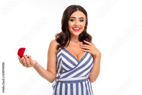 happy girl with brown hair in a dress with a neckline holds a box with an engagement ring on a white background