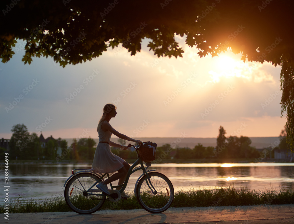 Obraz premium Happy woman riding a retro bike with a basket on the road near the lake on a sunset. Girl enjoying the magical view of the sun's rays of the falling sun at the end of the day