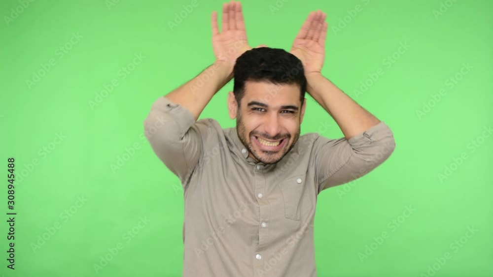 Joyful funny brunette man with bristle in shirt making bunny ears with hands over head, looking childish playful and carefree, entertaining, having fun. indoor studio shot, green background chroma key Joyful funny brunette man with bristle in shirt making bunny ears with hands over head, looking childish playful and carefree, entertaining, having fun. indoor studio shot, green background chroma key