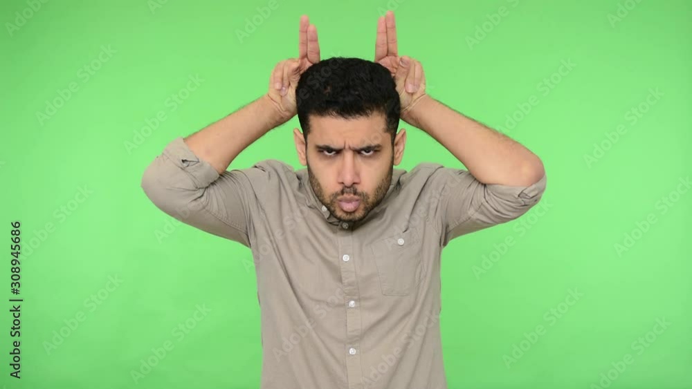 Aggressive brunette man with bristle in shirt making bull horn gesture on his head, looking with dangerous angry expression, threatening to attack. indoor studio shot, green background, chroma key Aggressive brunette man with bristle in shirt making bull horn gesture on his head, looking with dangerous angry expression, threatening to attack. indoor studio shot, green background, chroma key