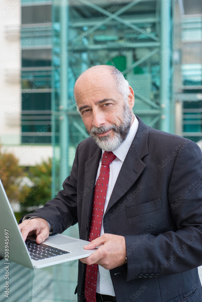 Positive confident grey haired businessman using laptop near office building. Elderly man in formal suit and tie walking outside in city. Business portrait concept Positive confident grey haired businessman using laptop near office building. Elderly man in formal suit and tie walking outside in city. Business portrait concept