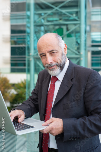 Positive confident grey haired businessman using laptop near office building. Elderly man in formal suit and tie walking outside in city. Business portrait concept