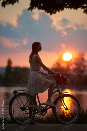 Romantic girl with a bike keeping her foot on a pedal is watching purple sunset at the lake, all colors of the sky, orange sun path, side view