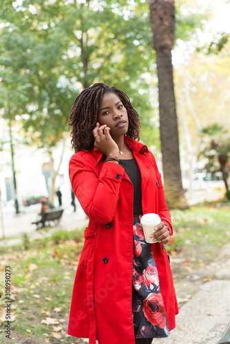 Woman talking by smartphone and looking at camera. Beautiful serious young African American woman holding coffee to go and talking by smartphone on street. Connection concept