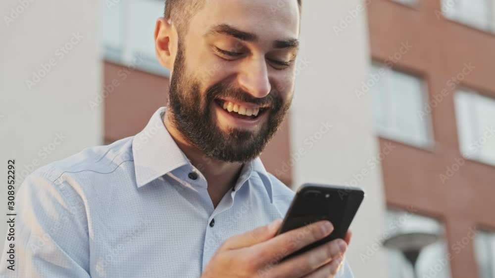 Close up view of Joyful bearded businessman using smartphone while sitting outdoors Close up view of Joyful bearded businessman using smartphone while sitting outdoors