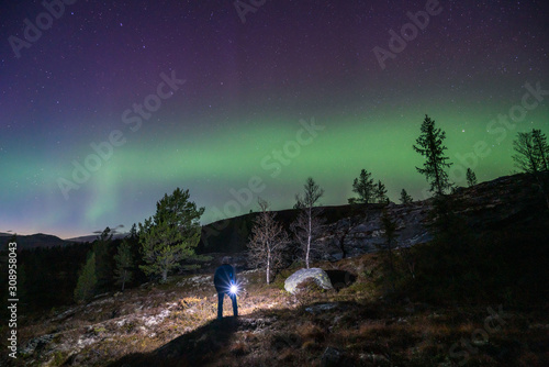 Northern lights/aurora borealis and starry sky from outdoors in the middle of the forest. Man stands in the foreground with flashlight. Galaxy, astronomy and nature phenomenon concept.