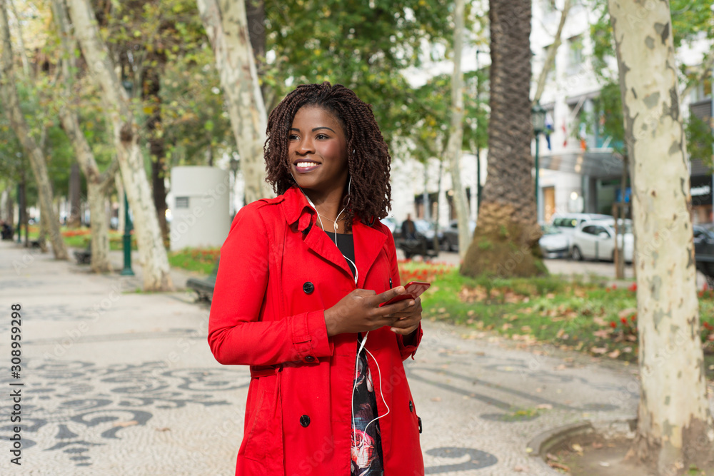 Content woman holding smartphone and walking on street. Cheerful young African American woman in headphones using cell phone outdoors. Technology concept Content woman holding smartphone and walking on street. Cheerful young African American woman in headphones using cell phone outdoors. Technology concept