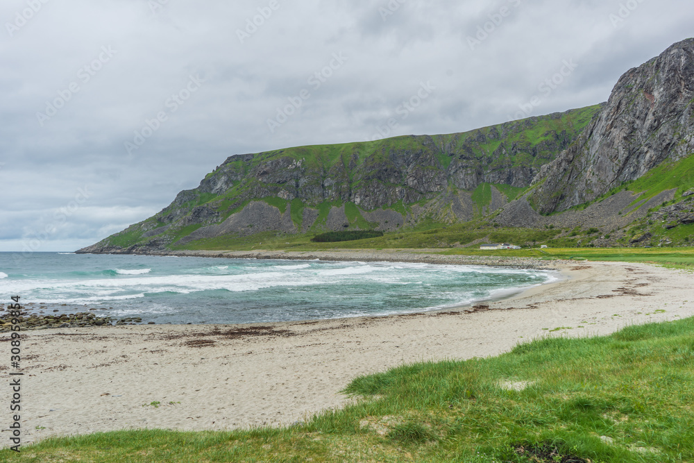A sand beach in clouded weater with green grass in the foreground and mountains in the background. Unstadt Beach, Lofoten Islands, Norway.