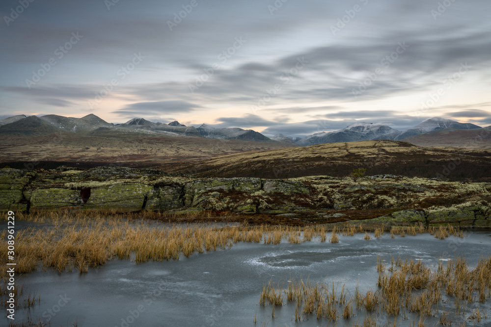 Tranquil scenery with frozen lake, snow covered mountains and fast moving skies over rondane national park in norway. Outdoors and blue hour theme. Tranquil scenery with frozen lake, snow covered mountains and fast moving skies over rondane national park in norway. Outdoors and blue hour theme.