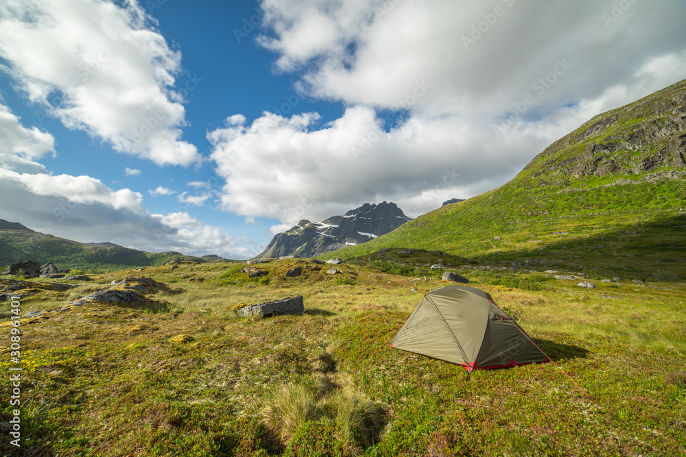 A green tent camouflaged in a green mountain valley. Above a blue sky with white cumulus clouds, Norway.