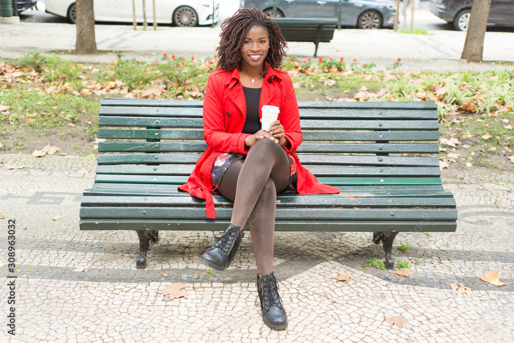 Woman with coffee to go smiling at camera. Full length view of cheerful young African American woman sitting on bench with paper cup. Hot drink concept Woman with coffee to go smiling at camera. Full length view of cheerful young African American woman sitting on bench with paper cup. Hot drink concept