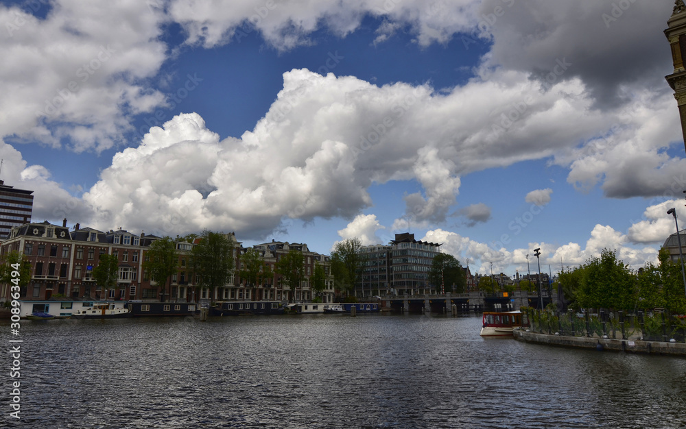 Amsterdam, Holland, August 2019. View of the Amstel River. On the shore boats house, behind the town. Blue sky with soft white clouds.