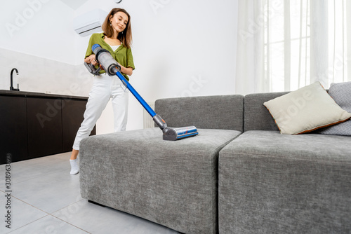 Young woman cleaning sofa with cordless vacuum cleaner in the modern white living room. Concept of easy cleaning with a wireless vacuum cleaner