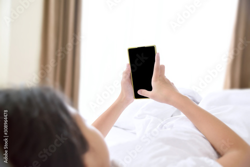 Concentrated young Asian girl looking at her smartphone lying on her bed in bedroom with clipping path for the inside. Technology and internet concept.