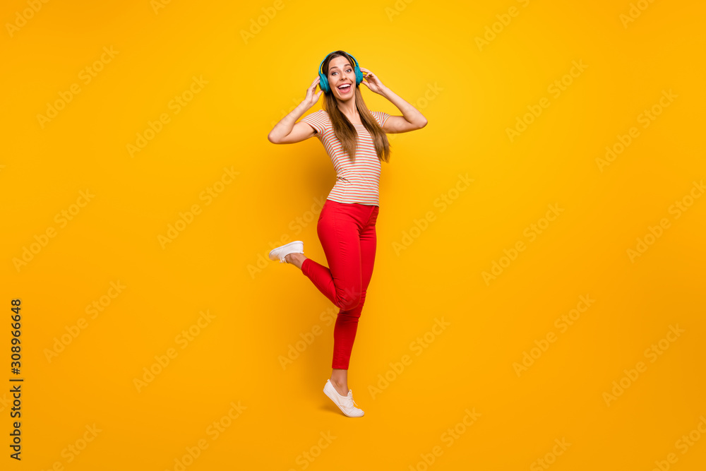 Full length photo of beautiful lady good mood walking down street listen music in cool wireless earphones wear casual striped t-shirt red trousers isolated yellow color background Full length photo of beautiful lady good mood walking down street listen music in cool wireless earphones wear casual striped t-shirt red trousers isolated yellow color background