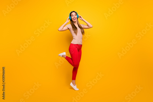 Full length photo of beautiful lady good mood walking down street listen music in cool wireless earphones wear casual striped t-shirt red trousers isolated yellow color background