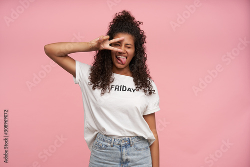 Shot of funny young curly long haired brunette woman with dark skin closing one eye and showing tongue to camera, folding victory sigh and keeping it near her face, isolated over pink background