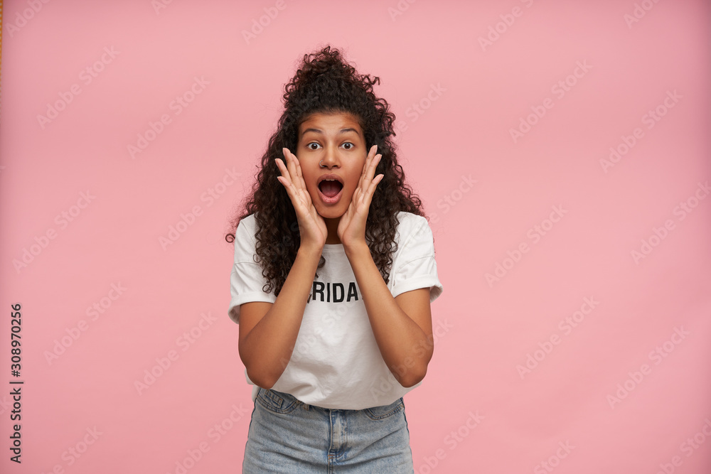 Astonished young curly dark skinned brunette female looking surprisedly to camera with raised eyebrows and holding face with hands, dressed in casual clothes while posing over pink background Astonished young curly dark skinned brunette female looking surprisedly to camera with raised eyebrows and holding face with hands, dressed in casual clothes while posing over pink background
