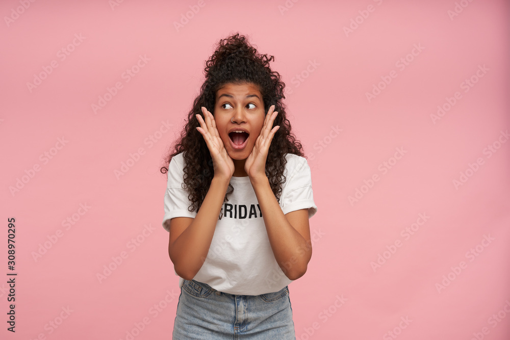 Surprised young curly long haired brunette woman with dark skin looking excited and keeping mouth wide opened, holding palms on her cheeks and rounding eyes amazedly over pink background Surprised young curly long haired brunette woman with dark skin looking excited and keeping mouth wide opened, holding palms on her cheeks and rounding eyes amazedly over pink background