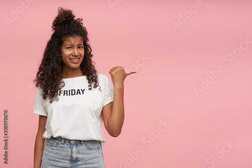 Displeased young pretty curly long haired brunette lady with dark skin frowning her face and showing aside with raised hand, wearing white t-shirt and blue jeans while posing over pink background