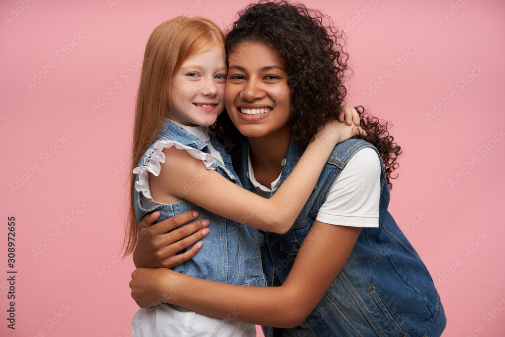Couple of happy lovely young ladies in jeans vests and white shirts embracing each other lovingly and looking cheerfully to camera with charming smiles, posing against pink background Couple of happy lovely young ladies in jeans vests and white shirts embracing each other lovingly and looking cheerfully to camera with charming smiles, posing against pink background