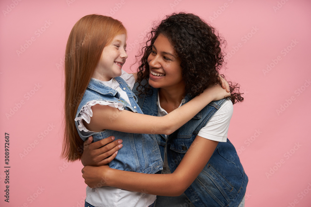 Indoor shot of two cheerful attractive young girls looking lovingly on each other and smiling sincerely, giving gentle hugs and having nice time together, isolated over pink background