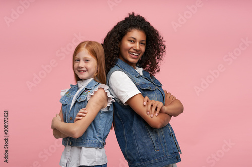 Positive attractive young ladies in casual clothes standing back to each other and crossing hands on chests while posing over pink background, looking cheerfully at camera and smiling widely