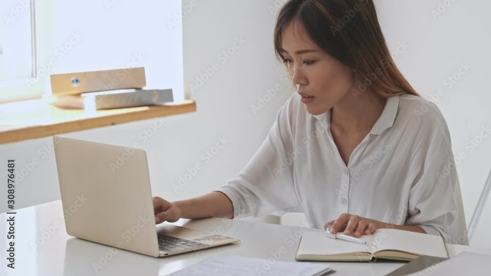 Serious asian business woman working with her notebook and making notes using a laptop while sitting by the table in the office Serious asian business woman working with her notebook and making notes using a laptop while sitting by the table in the office