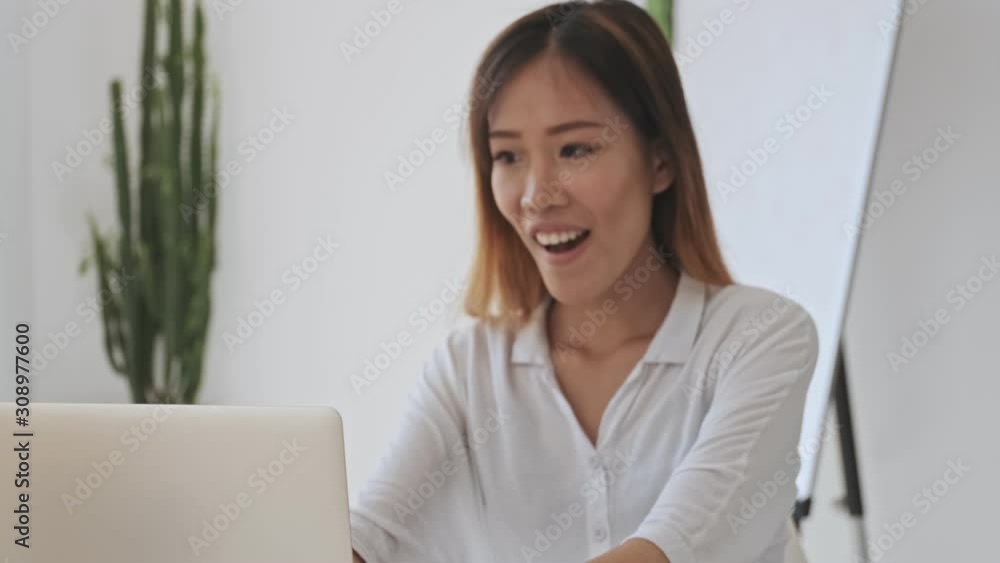 Happy asian business woman making a winner gesture while working with a laptop in the white office Happy asian business woman making a winner gesture while working with a laptop in the white office