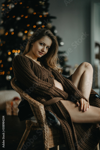 Pregnant woman sits in armchair against background of Christmas tree.