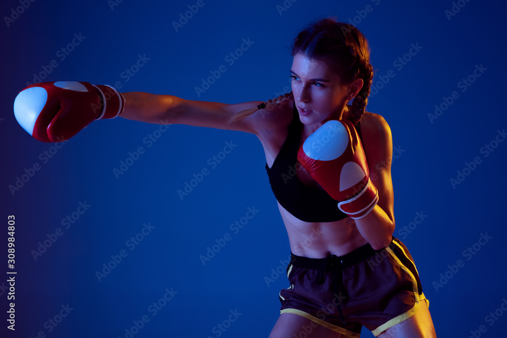 Fight. Fit caucasian woman in sportswear boxing on blue studio background in neon light. Novice female caucasian boxer working out and training. Sport, healthy lifestyle, movement concept.
