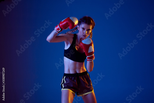 Fit caucasian woman in sportswear boxing on blue studio background in neon light. Novice female caucasian boxer working out and training. Sport, healthy lifestyle, movement concept. Copyspace.