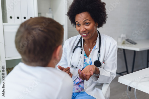 Smiling female afro american doctor general practitioner talks and amuses child before medical examination photo