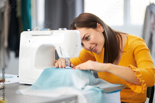 Smiling charming caucasian fashion designer sitting in her studio and sewing beautiful evening dress.