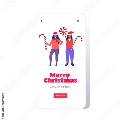 african american couple in santa claus hats holding sweetmeats candy cane and lollipop merry christmas happy new year winter holidays celebration concept smartphone screen online mobile app full