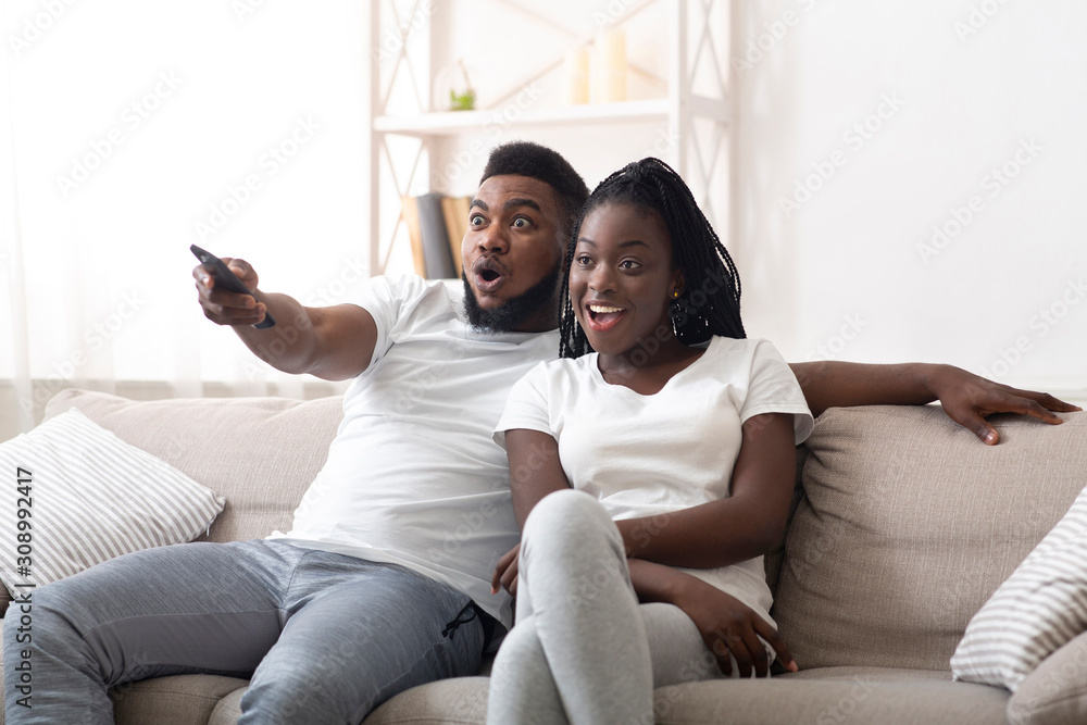 Excited afro couple watching tv together, sitting on couch at home