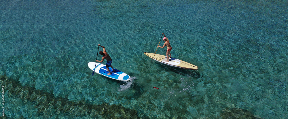 Aerial drone ultra wide photo of fit couple practising stand up paddle board or SUP board in tropical exotic sandy bay with turquoise sea
