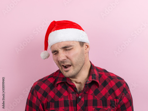 Emotions on the face, tired, holiday hangover, awareness. A man in a plaid rabbit and a Christmas red hat, on a pink background, copy space.