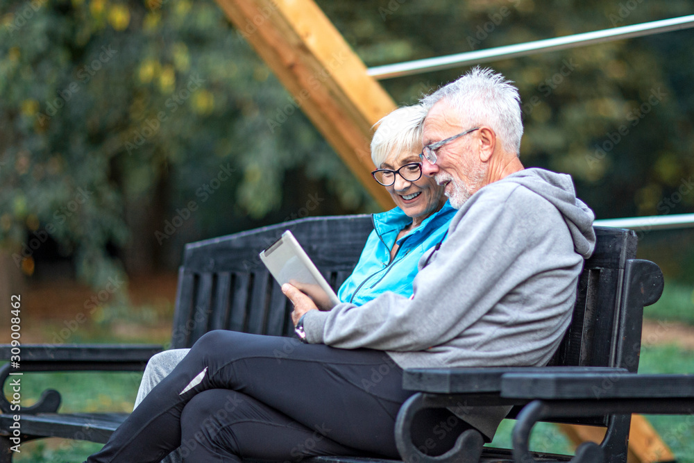 Mature couple sitting at banch in par and looking at tablet and having fun Mature couple sitting at banch in par and looking at tablet and having fun