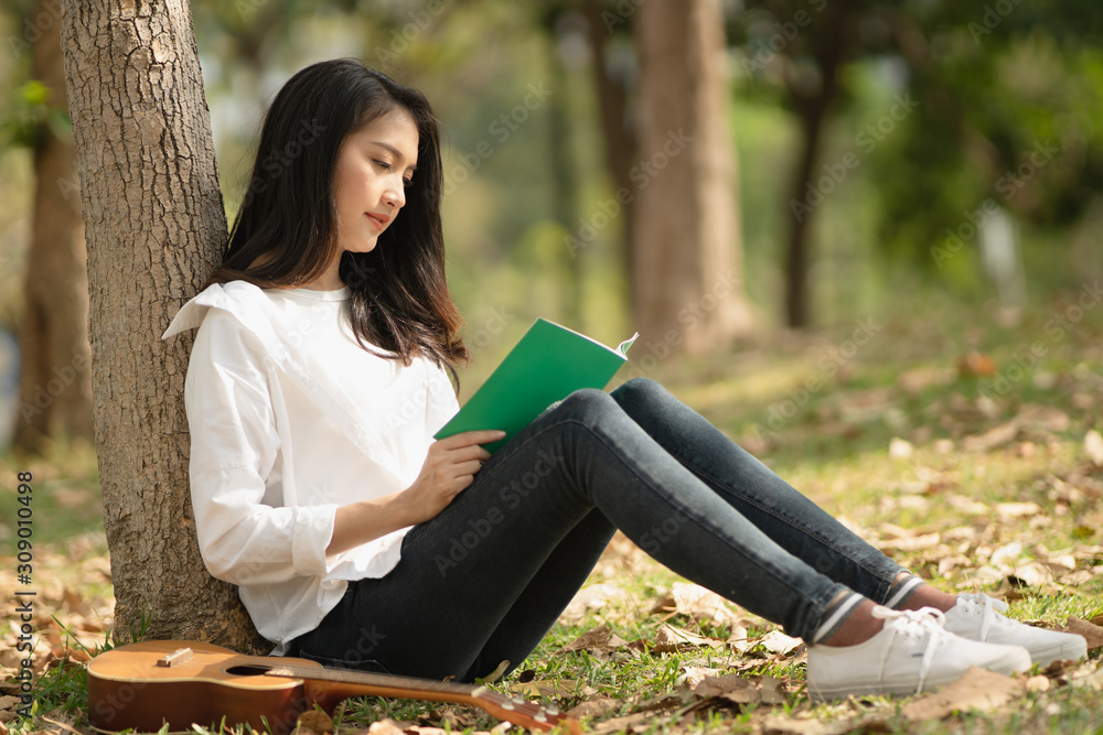 Side view of beautiful young Asian woman reading book at the park with ukulele at her side, concept leisure hobby time, reading is education, study by reading, music and book hobby, young people hobby Side view of beautiful young Asian woman reading book at the park with ukulele at her side, concept leisure hobby time, reading is education, study by reading, music and book hobby, young people hobby