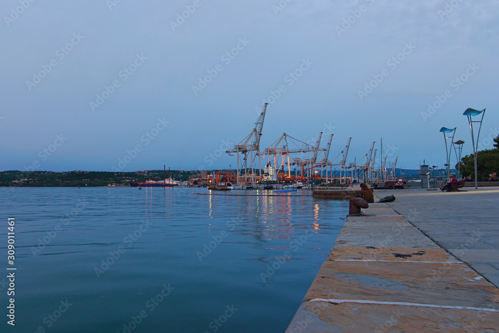 Koper, Slovenia-September 27, 2019: Scenic evening landscape view of Harbor of Koper. Many cranes in the background. People resting in the embankment near port. Romantic and peaceful scene Koper, Slovenia-September 27, 2019: Scenic evening landscape view of Harbor of Koper. Many cranes in the background. People resting in the embankment near port. Romantic and peaceful scene