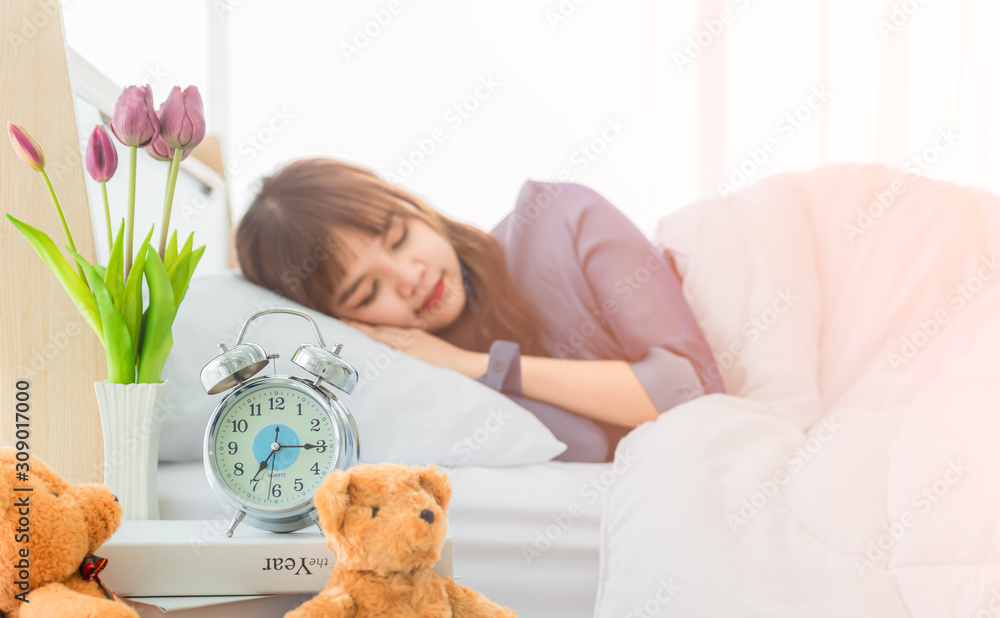Alarm clock on bedside table with flower vase and bear doll by young woman sleeping with happy in morning is background. Alarm clock on bedside table with flower vase and bear doll by young woman sleeping with happy in morning is background.