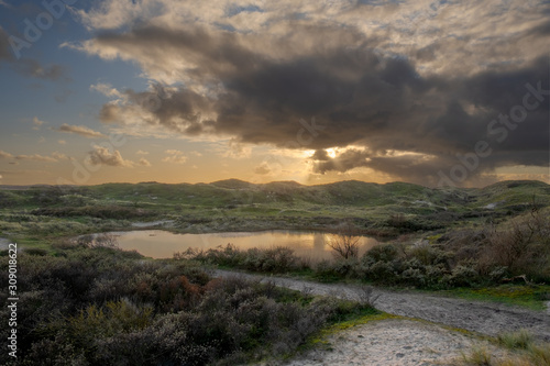 A small lake along a sandy path in the light of the sun from behind a dark cloud