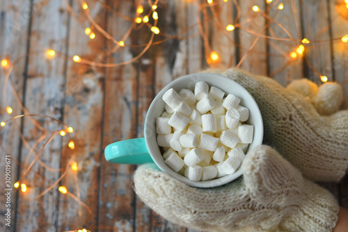 Woman's hand in white mittens holding mug of Christmas cocoa with marshmallows on wooden background with bokeh New Year's lights. . Drink For Winter Season. Comfort Food Concept