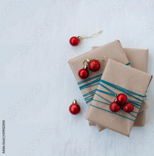 Three gifts wrapped in craft paper with bright red Christmas balls in the form of balls. Gifts are rewound with blue thread. Gifts lie on white wooden background. There is a place for greeting text.