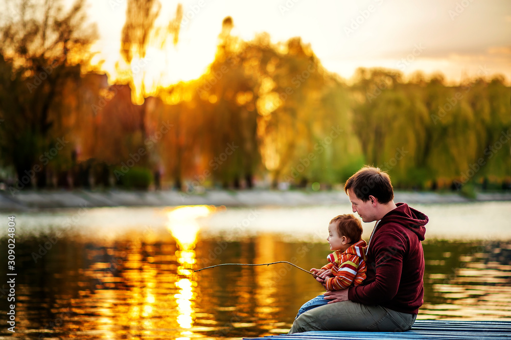 Father and 2 years old child play fishing on bank of lake in sunset time. Golden in sun light trees and water around. Father and daughter son fun together. Lesson of fishing in evening time. Weekend Father and 2 years old child play fishing on bank of lake in sunset time. Golden in sun light trees and water around. Father and daughter son fun together. Lesson of fishing in evening time. Weekend