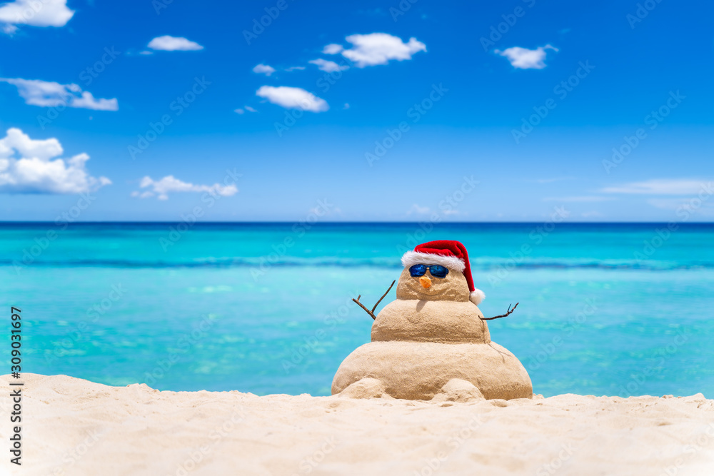 Obraz premium Smiling sandy snowman with red santa hat on the caribbean beach with beautiful sky . Holiday concept for New Year and Christmas Cards