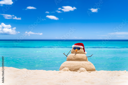 Smiling sandy snowman with red santa hat on the caribbean beach with beautiful sky . Holiday concept for New Year and Christmas Cards