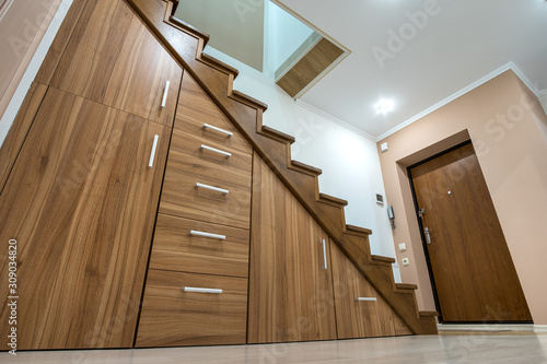Modern architecture interior with luxury hallway with glossy wooden stairs in multi-storey house. Custom built pullout cabinets on glides in slots under stairs. Use of space for storage.