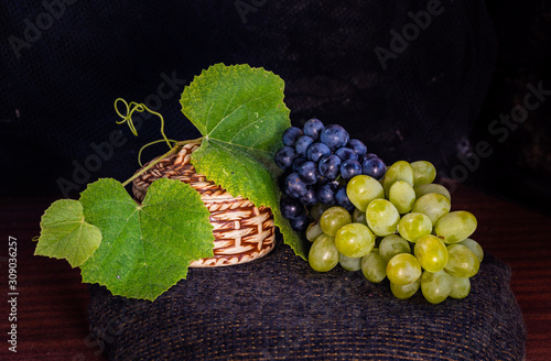 white and black clusters of grapes with green leaves lie next to a wicker small basket